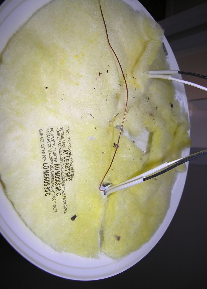 The underside of a round white light fixture, meaning the side that is hidden aginst the ceiling. 
	We see black, white, and bare wires, yellow fiberglas thermal insulation, and a warning, 
	'For supply connections use only wire suitable for at least 90 degree C,' stated 
	in English, French, and Spanish.