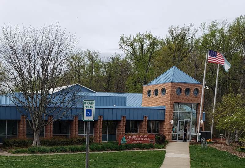 Aquatic-fitness center: foreground, grassy lawn with one concrete walk/ramp going gradually up to the front door and 
another horizontally left-to-right along the building. To the right of the 
building we see bicyle racks and the railings at the top of the (off-camera) stairs
that descend to the right. At the left end of the visible building we see a 
young tree, behind the horizontal walkway, before the plantings right in front of 
the building. The main part of the building is red brick with floor-to-ceiling 
windows, and a blue roof. The main entrance is a two story tower, with glass doors, 
plus small glass windows above, and a blue roof. In front, to the right
of the main entrance, we see two flagpoles. On one a U.S. flag waves; on the other,
Greenbelt's flag. Behind the building we see green trees, and above them, pale blue-gray sky.