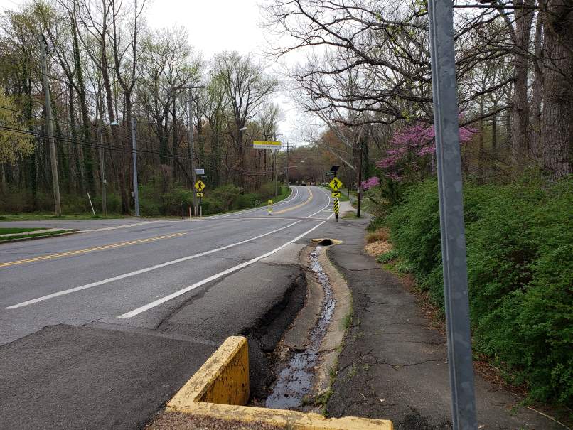 A curving stretch of Crescent Road. On the immediate right is 
a galvanized pole. We see a culvert with water flowing; a black macadam footpath 
to its right; a marked bicycle lane; two marked lanes, curving toward the back 
of the picture. On either side we see bushes and trees. In the middle distance we 
see two 'Walk'signs attached to structures. One structure has a small photocell array on 
top. (They're too far--meaning too small in the picture--for viewers to make out the red
'push to walk' buttons.)